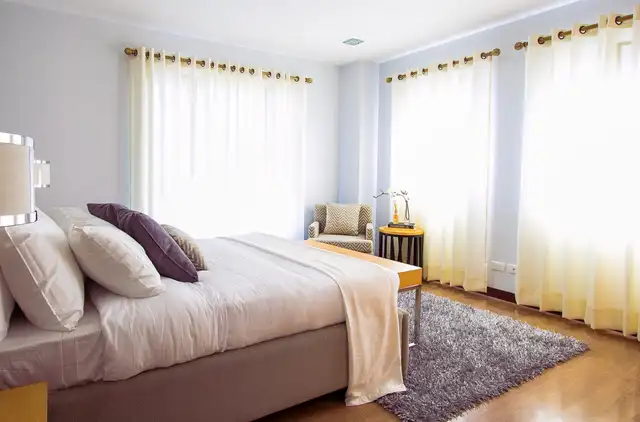 A bedroom with a made bed, a grey rug and long white curtains