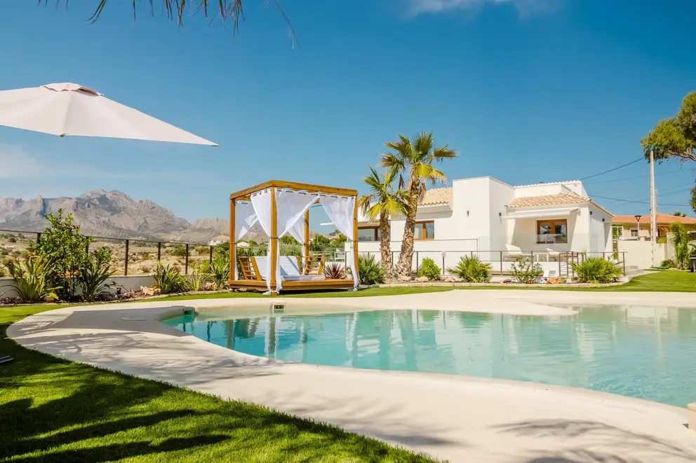 A pool beside a white house, with a pergola and mountains behind