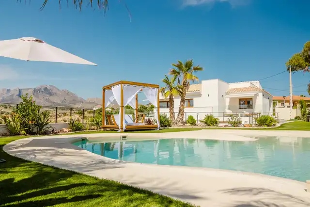 A pool beside a white house, with a pergola and mountains behind