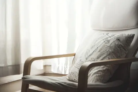 A wooden armchair with a leaf-pattern cushion by a curtained window
