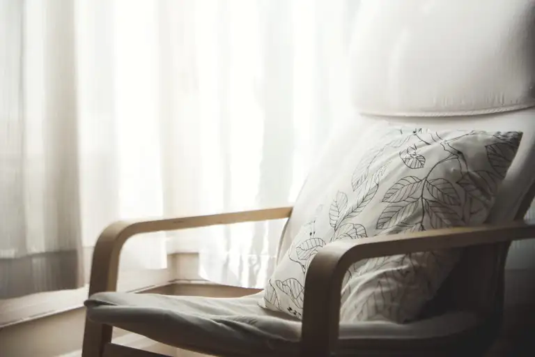 A wooden armchair with a leaf-pattern cushion by a curtained window